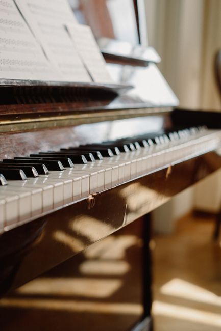 A close-up view of a polished wooden upright piano with visible black and white keys, situated indoors near a window with sheer curtains allowing natural light to illuminate the scene. A sheet of musi